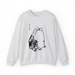 May include: A light gray sweatshirt with a black and white illustration of a frog on the front. The frog is sitting on its hind legs with its front legs outstretched. The illustration is in a Japanese style and includes the Japanese characters "蛙" (frog) and a red circle.