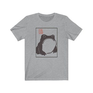 May include: A grey t-shirt with a black and white illustration of a frog in a square frame. The frog is looking up and has a large, round body. There is a red stamp in the top left corner of the frame.
