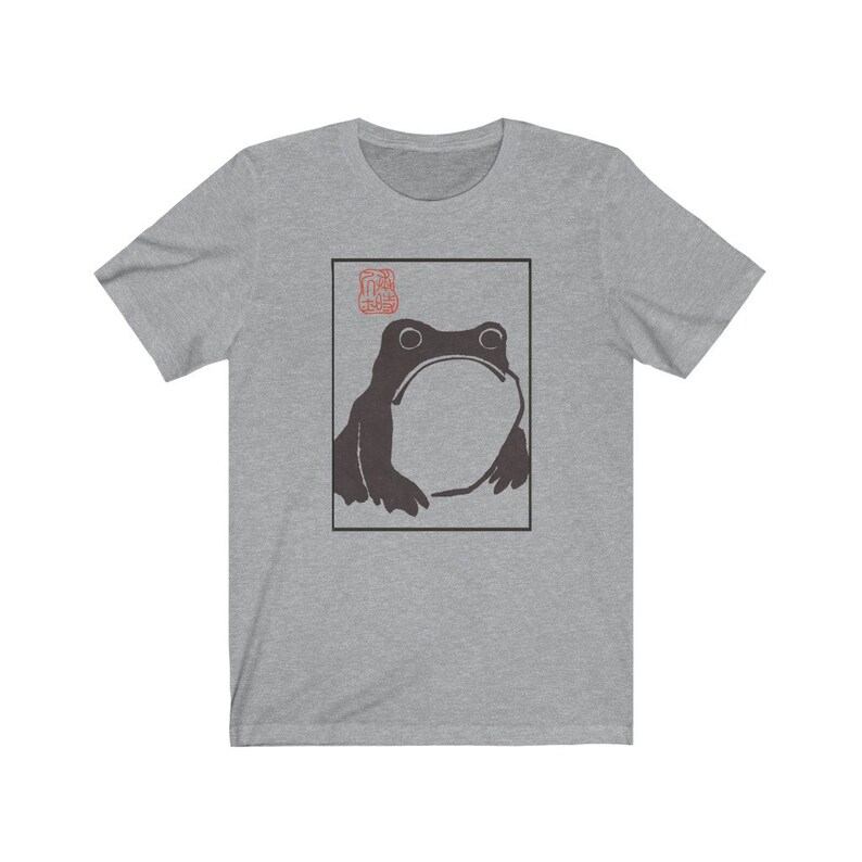 May include: A grey t-shirt with a black and white illustration of a frog in a square frame. The frog is looking up and has a large, round body. There is a red stamp in the top left corner of the frame.