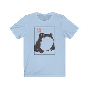 May include: Light blue t-shirt with a black and white illustration of a frog in a square frame. The frog is looking up and has a large, round belly. There is a red stamp in the top left corner of the frame.