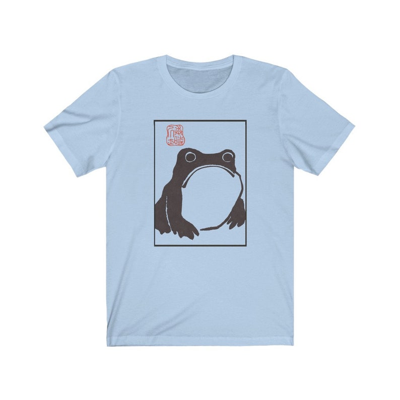 May include: Light blue t-shirt with a black and white illustration of a frog in a square frame. The frog is looking up and has a large, round belly. There is a red stamp in the top left corner of the frame.