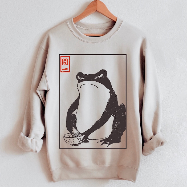 May include: A beige sweatshirt with a black and white illustration of a frog holding a bowl. The frog is sitting in a square frame with the Japanese character "開" in red in the top left corner.