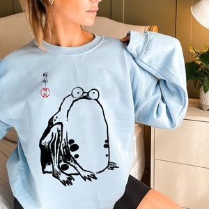 May include: A light blue sweatshirt with a black and white illustration of a frog. The frog is sitting on its hind legs with its front legs outstretched. The sweatshirt has Japanese characters printed on the front.
