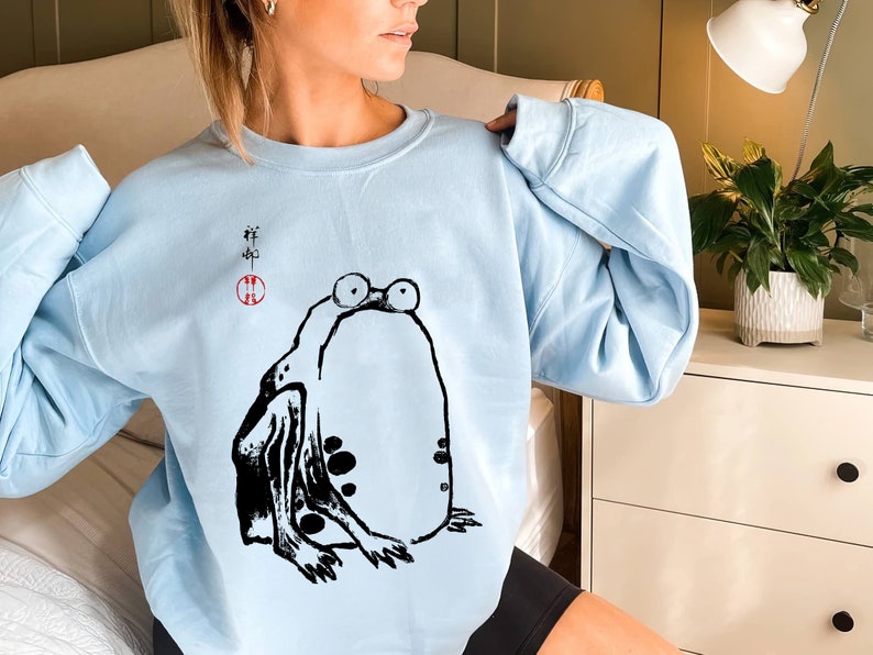 May include: A light blue sweatshirt with a black and white illustration of a frog. The frog is sitting on its hind legs with its front legs outstretched. The sweatshirt has Japanese characters printed on the front.