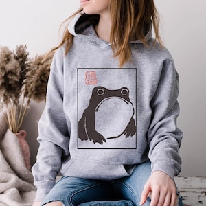 May include: A grey hoodie featuring a black and white illustration of a frog within a square frame. The frog is looking upwards and has a large mouth. A red stamp is located in the top left corner of the frame.