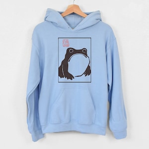 May include: A light blue hoodie featuring a black and white illustration of a frog within a square frame. The frog is looking upwards and has a large, round body. A red stamp is visible in the top left corner of the frame.
