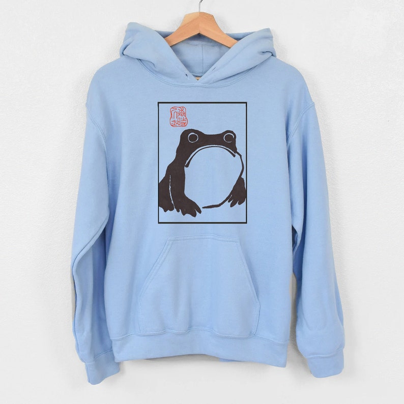 May include: A light blue hoodie featuring a black and white illustration of a frog within a square frame. The frog is looking upwards and has a large, round body. A red stamp is visible in the top left corner of the frame.