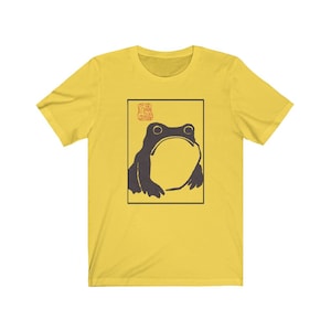 May include: A yellow t-shirt with a black and white illustration of a frog in a square frame. The frog is looking up and has a large mouth. The frame has a red stamp with Japanese characters in the top left corner.
