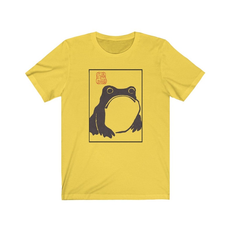 May include: A yellow t-shirt with a black and white illustration of a frog in a square frame. The frog is looking up and has a large mouth. The frame has a red stamp with Japanese characters in the top left corner.