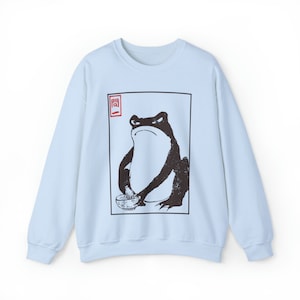 May include: Light blue sweatshirt with a black and white illustration of a frog holding a bowl. The illustration is framed in a black square with a red square in the top left corner that contains a Japanese character.
