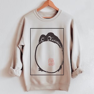 Puede incluir: Una sudadera beige con un gráfico en blanco y negro de un círculo con un sello rojo en su interior. El sello contiene caracteres japoneses.