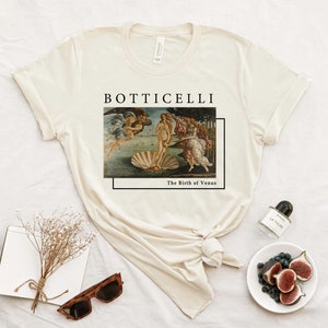 May include: A beige t-shirt with a graphic print of Sandro Botticelli's "The Birth of Venus" painting. The text "Botticelli" and "The Birth of Venus" is printed below the image.