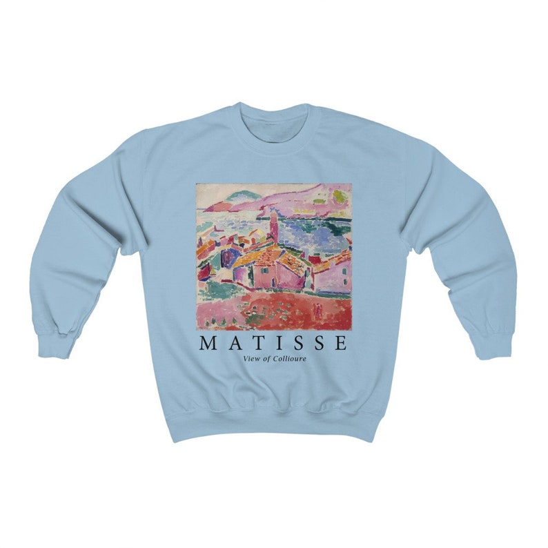 May include: Pale blue sweatshirt with a vibrant print of a Matisse painting of a coastal scene. The artwork shows buildings with red roofs, a sea, and the text "MATISSE View of Collioure" in black.