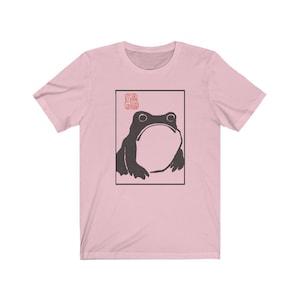 May include: A pink t-shirt with a black and white illustration of a frog in a square frame. The frog is looking up and has a large, round belly. The word "蛙" is printed in red above the frog.