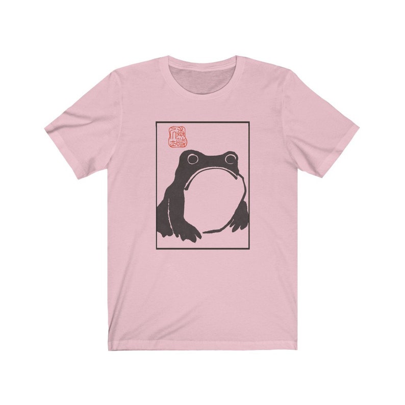 May include: A pink t-shirt with a black and white illustration of a frog in a square frame. The frog is looking up and has a large, round belly. The word "蛙" is printed in red above the frog.