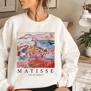 May include: White sweatshirt with a vibrant print of a Matisse landscape painting. The artwork shows a coastal scene with red-roofed buildings, a blue sea, and a pink mountain range. The word "MATISSE" is printed below the artwork, with "View of Collioure" underneath.