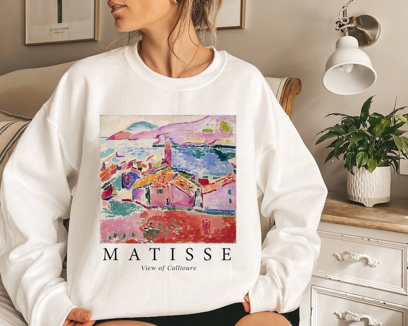 May include: White sweatshirt with a vibrant print of a Matisse landscape painting. The artwork shows a coastal scene with red-roofed buildings, a blue sea, and a pink mountain range. The word "MATISSE" is printed below the artwork, with "View of Collioure" underneath.