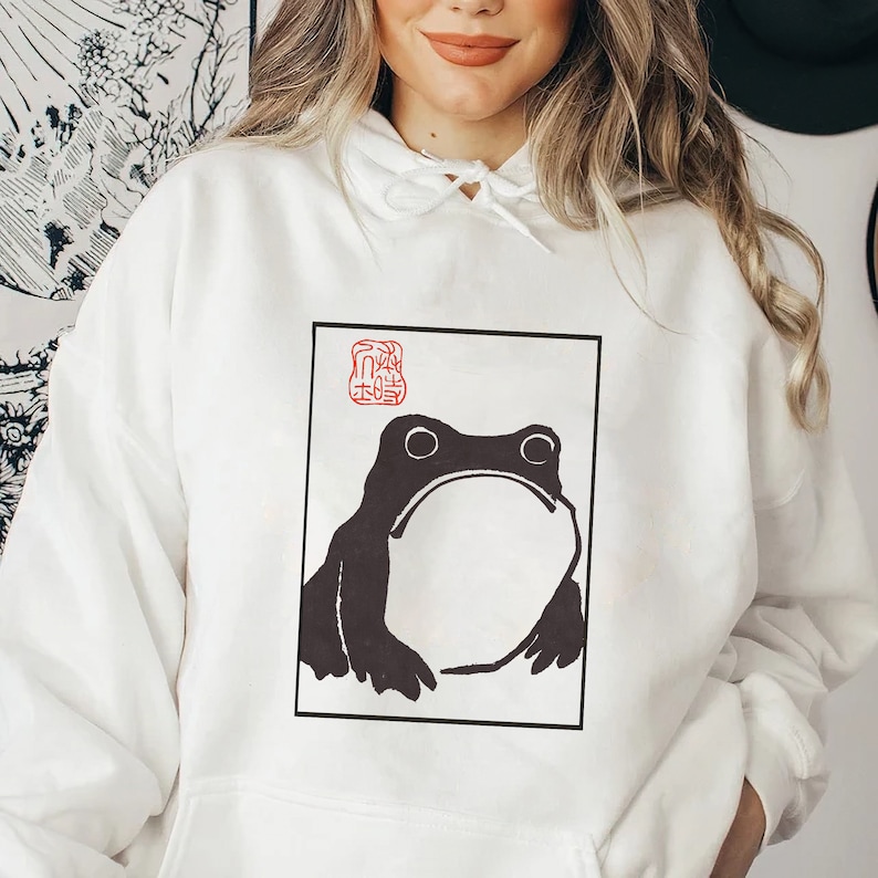 May include: White hoodie featuring a black and white illustration of a frog within a square frame. The frog is facing forward with its mouth open. The frame includes a red stamp with Japanese characters in the top left corner.