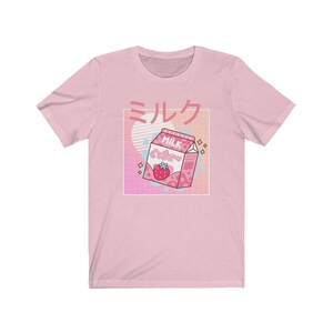 Op de afbeelding: Lichtroze t-shirt met een afbeelding van een roze aardbeienmelkpak en Japanse tekst. Het ontwerp bevat een witte vierkant, een roze en blauwe gradi&euml;nt en een witte cirkel.