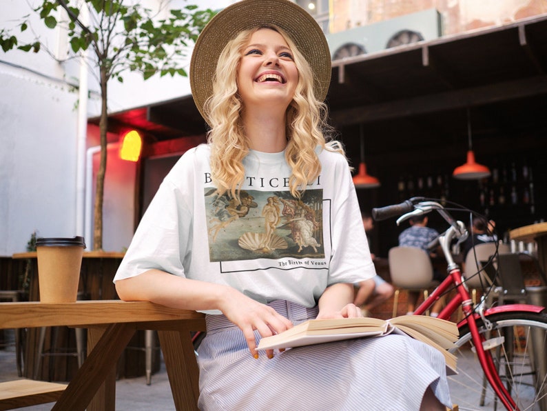 May include: A white t-shirt with a print of the painting "The Birth of Venus" by Sandro Botticelli. The shirt has a crew neck and short sleeves. The model is wearing a straw hat and smiling.