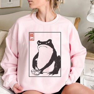 May include: A pink sweatshirt with a black and white graphic of a frog holding a bowl. The graphic is in a white square with a red square in the top left corner that says "開".
