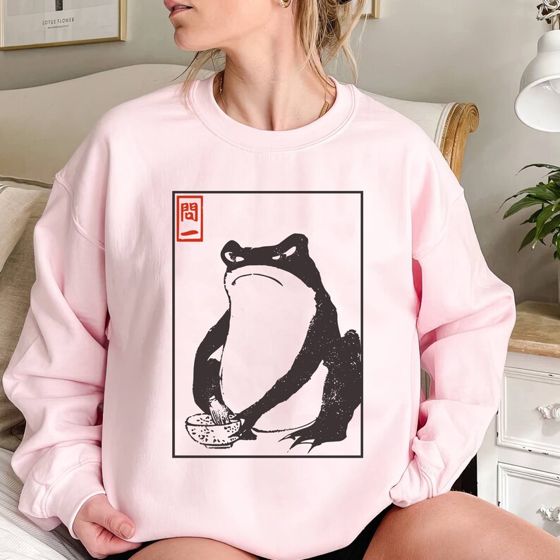 May include: A pink sweatshirt with a black and white graphic of a frog holding a bowl. The graphic is in a white square with a red square in the top left corner that says "開".