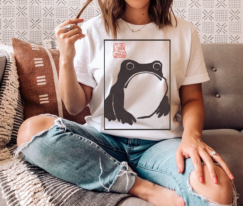 May include: A white t-shirt with a black and white illustration of a frog in a square frame. The frog is looking directly at the viewer. The shirt has a red stamp with Japanese characters in the top left corner of the frame.