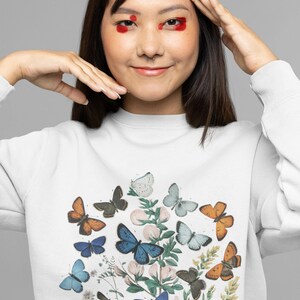 Puede incluir: Sudadera blanca con estampado de mariposas de colores. Las mariposas est&aacute;n dispuestas en un grupo en la parte delantera de la camiseta. Las mariposas son de tonos azules, naranjas y blancos.