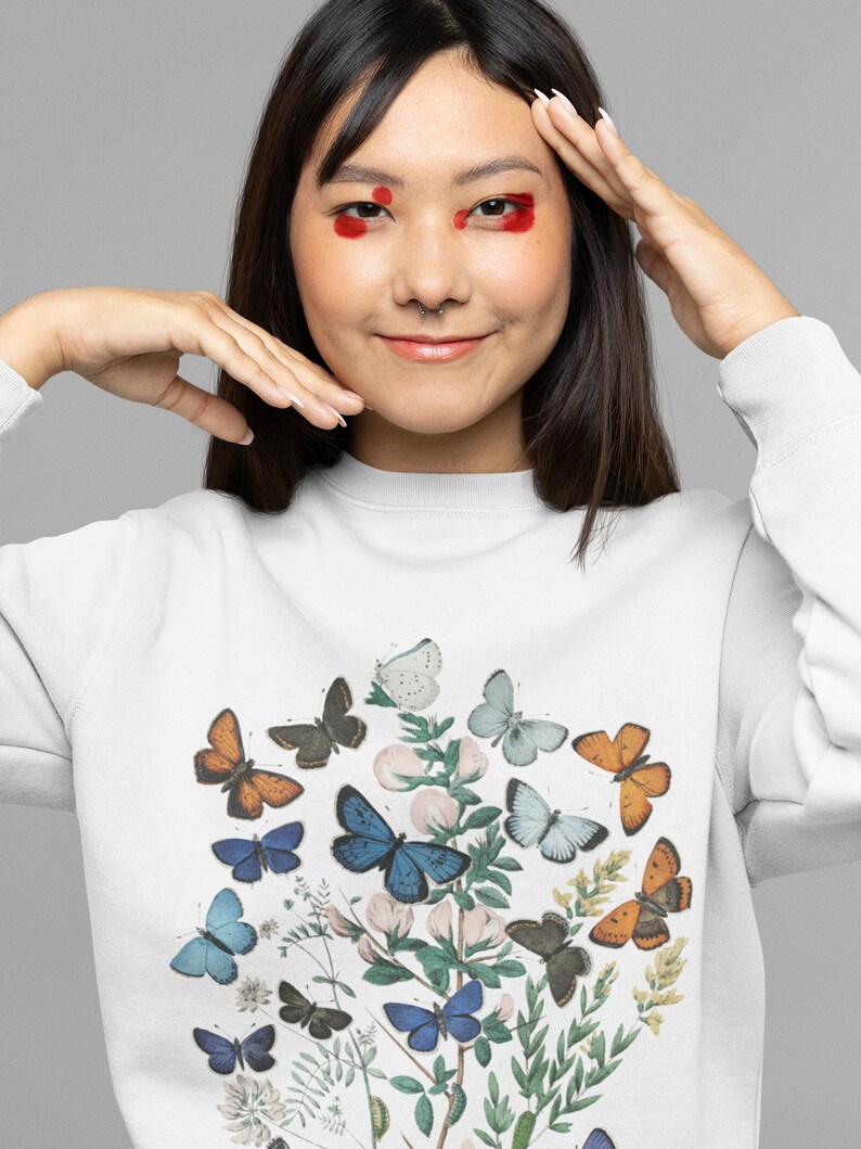 Puede incluir: Sudadera blanca con estampado de mariposas de colores. Las mariposas est&aacute;n dispuestas en un grupo en la parte delantera de la camiseta. Las mariposas son de tonos azules, naranjas y blancos.