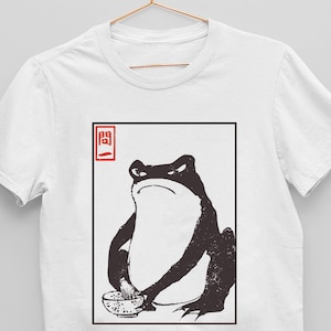 以下が含まれることがあります： 白いTシャツに、麺の入ったボウルを持ったカエルの白黒イラストが描かれています。イラストは黒い正方形で囲まれており、左上に赤い正方形の中に「開」という日本語の文字が書かれています。