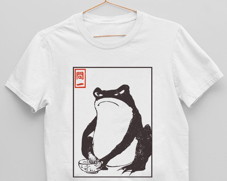 以下が含まれることがあります： 白いTシャツに、麺の入ったボウルを持ったカエルの白黒イラストが描かれています。イラストは黒い正方形で囲まれており、左上に赤い正方形の中に「開」という日本語の文字が書かれています。