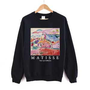 May include: A black sweatshirt displaying a vibrant print of a Matisse painting. The artwork shows a coastal scene with red-roofed buildings and a blue sea. The text "MATISSE View of Collioure" is printed in white below the image.