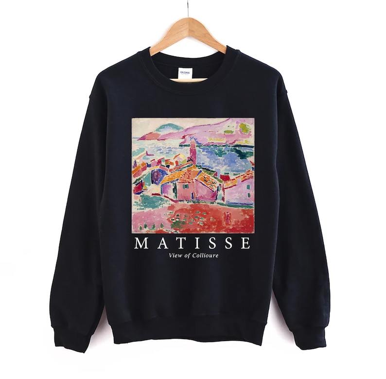 May include: A black sweatshirt displaying a vibrant print of a Matisse painting. The artwork shows a coastal scene with red-roofed buildings and a blue sea. The text "MATISSE View of Collioure" is printed in white below the image.