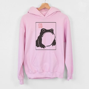 May include: Pink hoodie featuring a black and white illustration of a frog within a square frame. The frog is looking upwards and has a large mouth. A red stamp is positioned above the frog.
