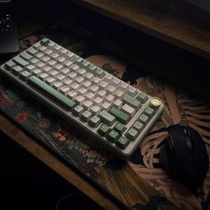 May include: A mechanical keyboard with white and green keycaps, a gold-colored knob, and a matching green frame. The keyboard sits on a patterned mousepad with a black mouse to the right. The keyboard is a modern computer accessory.
