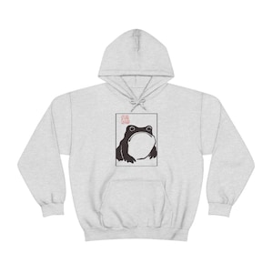 May include: A grey hooded sweatshirt with a black and white illustration of a frog on the front. The frog is looking up and has a large mouth. The illustration is surrounded by a white border. The word "Frog" is written in red above the frog.