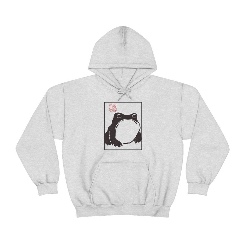 May include: A grey hooded sweatshirt with a black and white illustration of a frog on the front. The frog is looking up and has a large mouth. The illustration is surrounded by a white border. The word "Frog" is written in red above the frog.