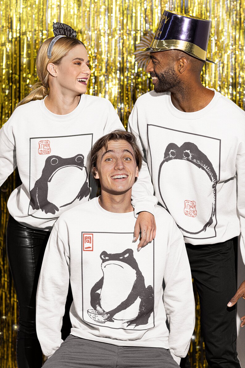 May include: Three people wearing white sweatshirts with black and white frog illustrations. The sweatshirts have red Japanese characters on them. The people are standing in front of a gold background.