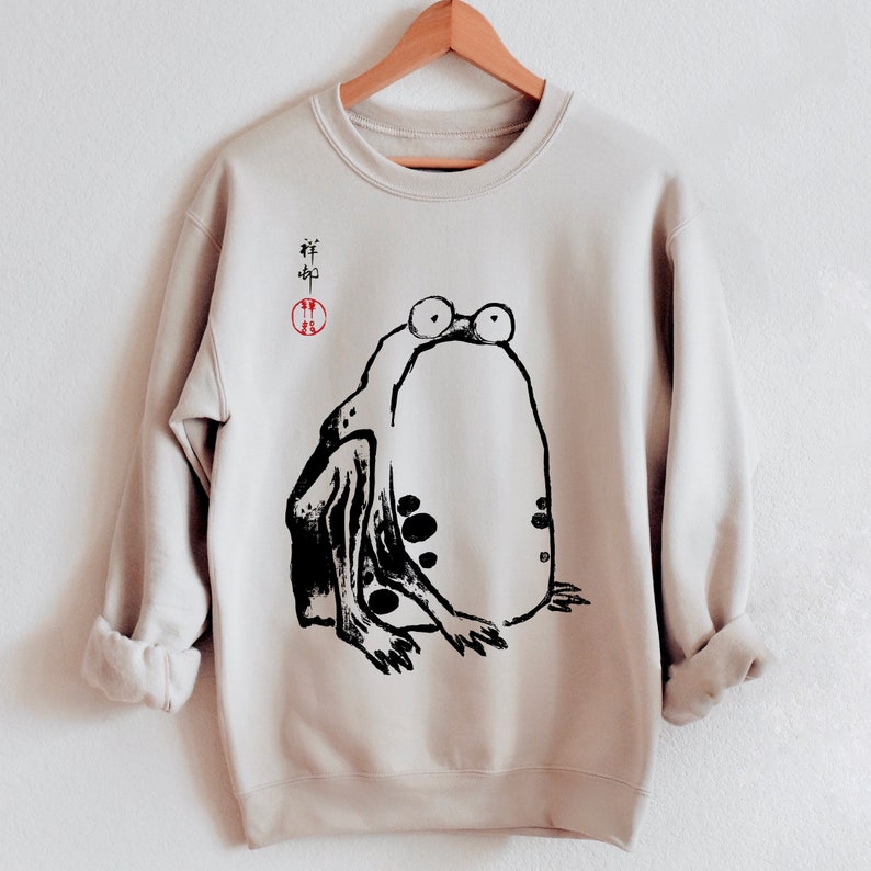 May include: A beige sweatshirt with a black and white illustration of a frog on the front. The frog is sitting on its hind legs with its front legs outstretched. The sweatshirt has a red circle with Chinese characters in it on the left side of the chest.