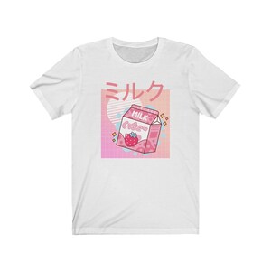 Op de afbeelding: Wit T-shirt met een roze en witte afbeelding van een aardbeienmelkpak en Japanse tekst. Het ontwerp bevat een roze raster, een lichtblauwe cirkel en meer Japanse tekst. Een casual kledingstuk.