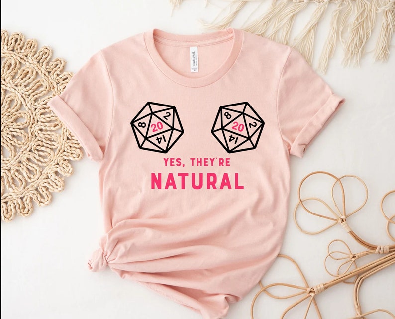 May include: Pink t-shirt with a graphic of two dice with the number 20 showing on each die. The text "YES, THEY'RE NATURAL" is printed below the dice.