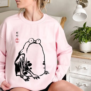 May include: Pink sweatshirt with a black and white illustration of a frog. The frog is sitting on its hind legs with its front legs outstretched. The text "#" is written in black above the frog.