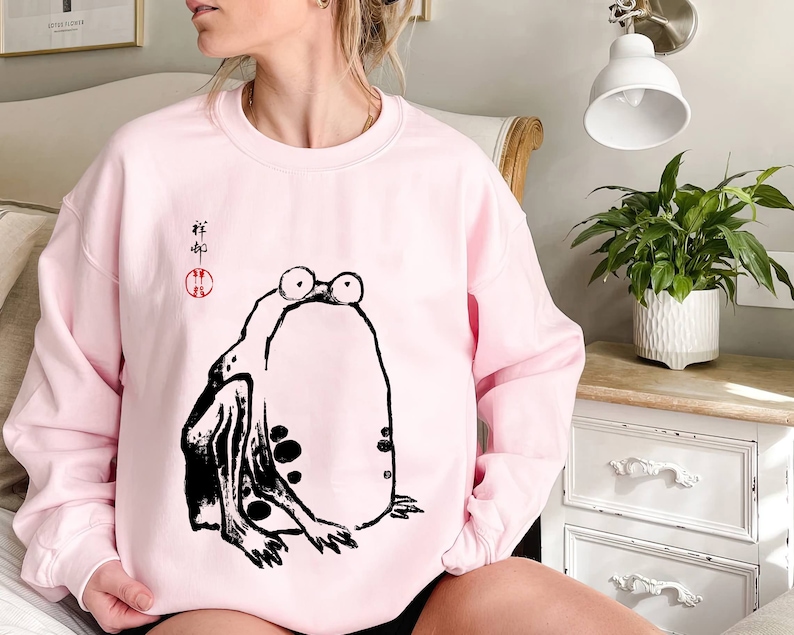 May include: Pink sweatshirt with a black and white illustration of a frog. The frog is sitting on its hind legs with its front legs outstretched. The text "#" is written in black above the frog.
