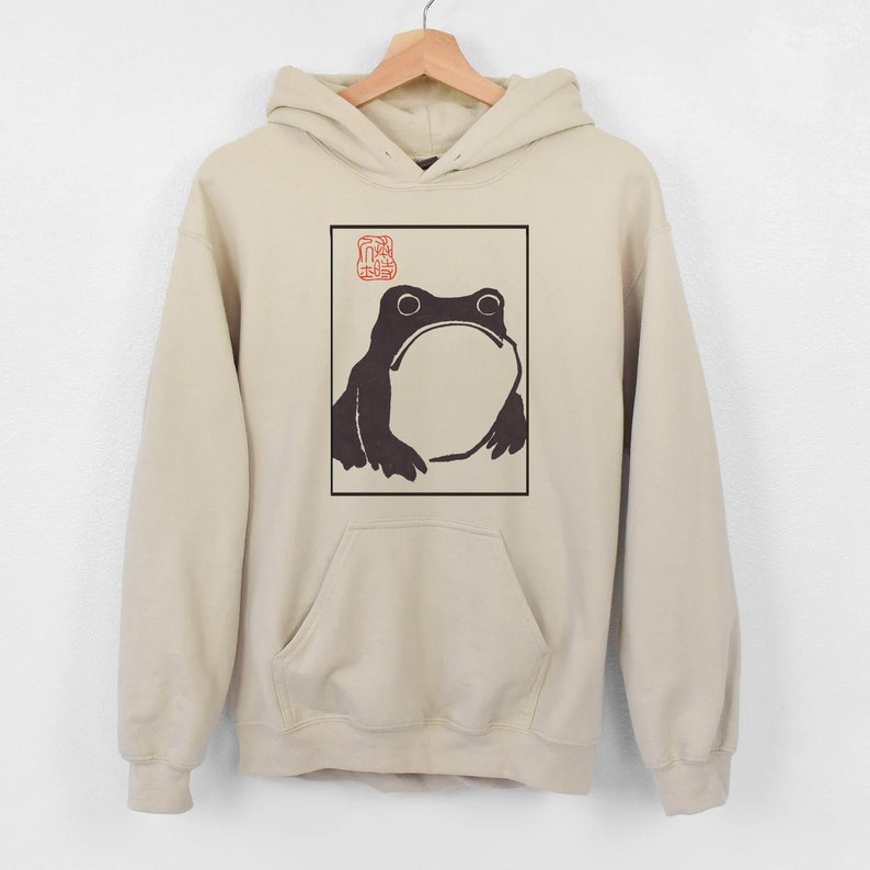 May include: A beige hoodie featuring a black and white illustration of a frog within a frame. The frog is looking upwards and has a large belly. A red stamp is positioned above the frog.