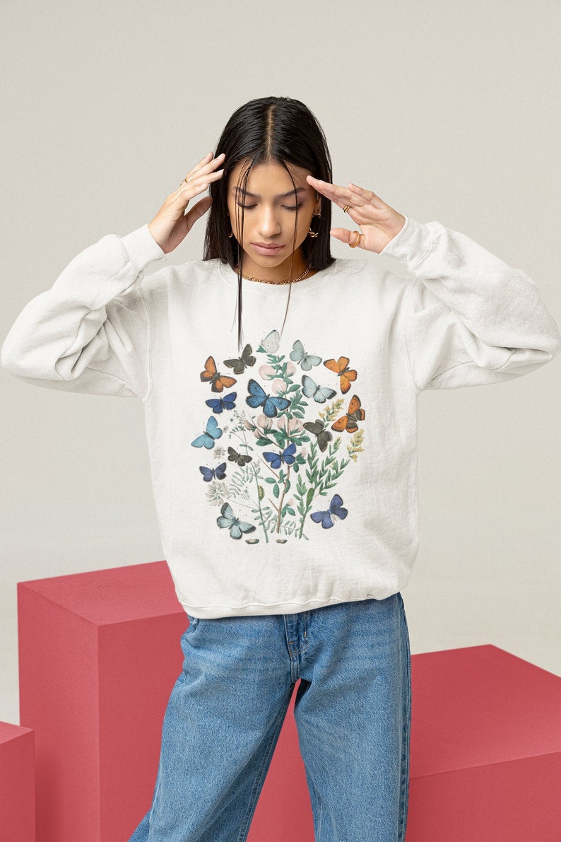 Puede incluir: Una sudadera blanca con un estampado de mariposas de colores. Las mariposas est&aacute;n agrupadas en el frente de la sudadera, con algunas mariposas volando hacia un lado. La sudadera la lleva puesta una persona con el pelo largo y oscuro.