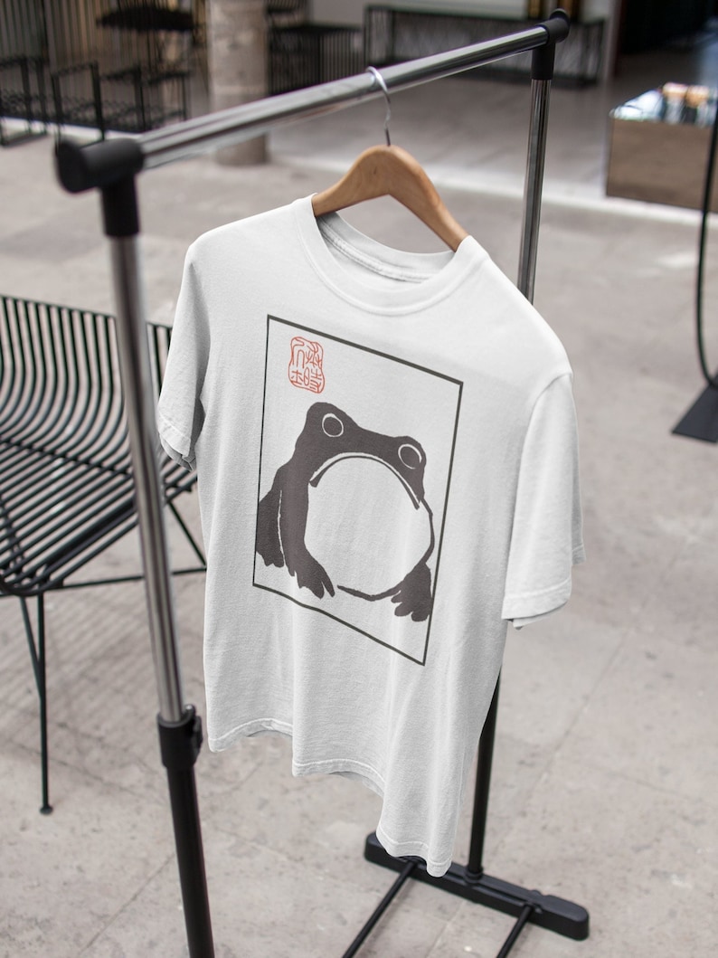 May include: White t-shirt with a black and white illustration of a frog in a square frame. The frame has a red stamp with Japanese characters in the top left corner.