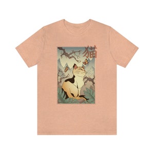May include: A peach-colored t-shirt with a graphic print of a cat sitting in a Japanese garden setting. The cat is looking up at butterflies and the text "猫" is printed in red above the cat.