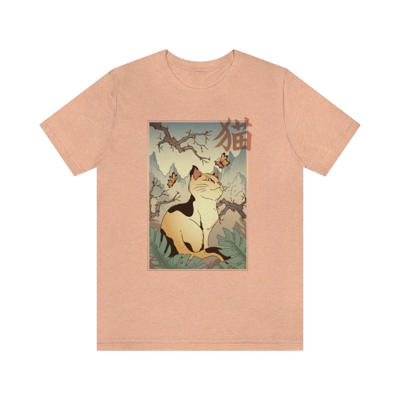 May include: A peach-colored t-shirt with a graphic print of a cat sitting in a Japanese garden setting. The cat is looking up at butterflies and the text "猫" is printed in red above the cat.