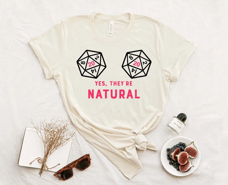 May include: A white t-shirt with a graphic of two dice with the number 20 showing. The text "YES, THEY'RE NATURAL" is printed below the dice.