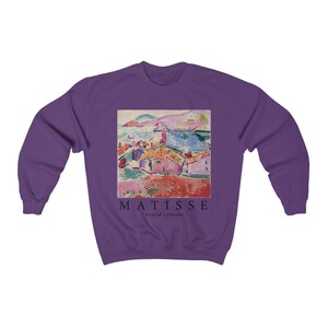 May include: A purple sweatshirt with a printed image of a coastal landscape painting. The artwork shows a town with red-tiled roofs, the sea, and mountains. The text "MATISSE View of Collioure" is printed below the painting in black.